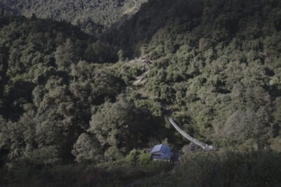 view from above, hikers on the trail, a suspension bridge, a small hut with a blue roof, rolling hills, a dense forest