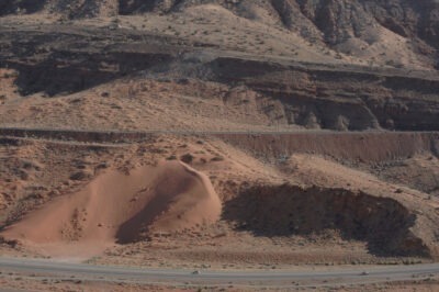 the side of a mountain and a road at the bottom of it, cars driving by, red sand, outside of Moab, Utah